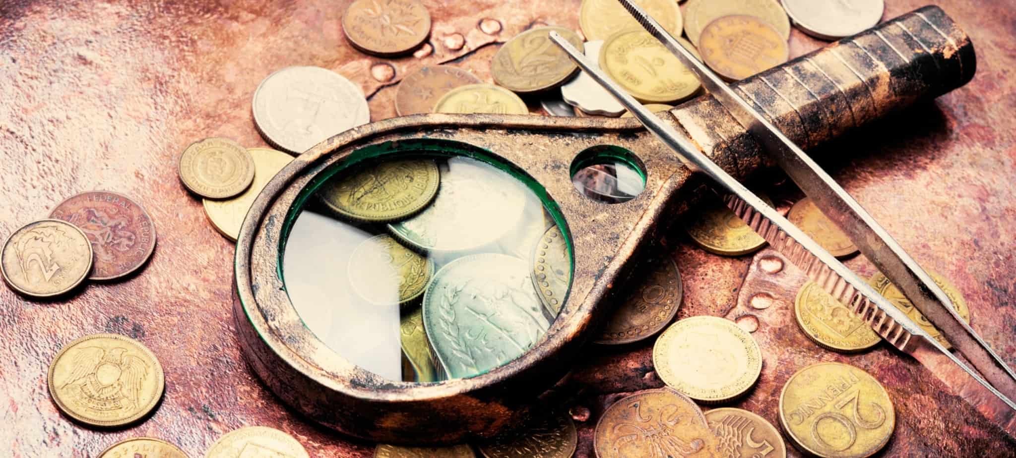 Várias moedas com aspeto antigo sobre uma mesa de madeira. Em cima uma lupa e uma pinça