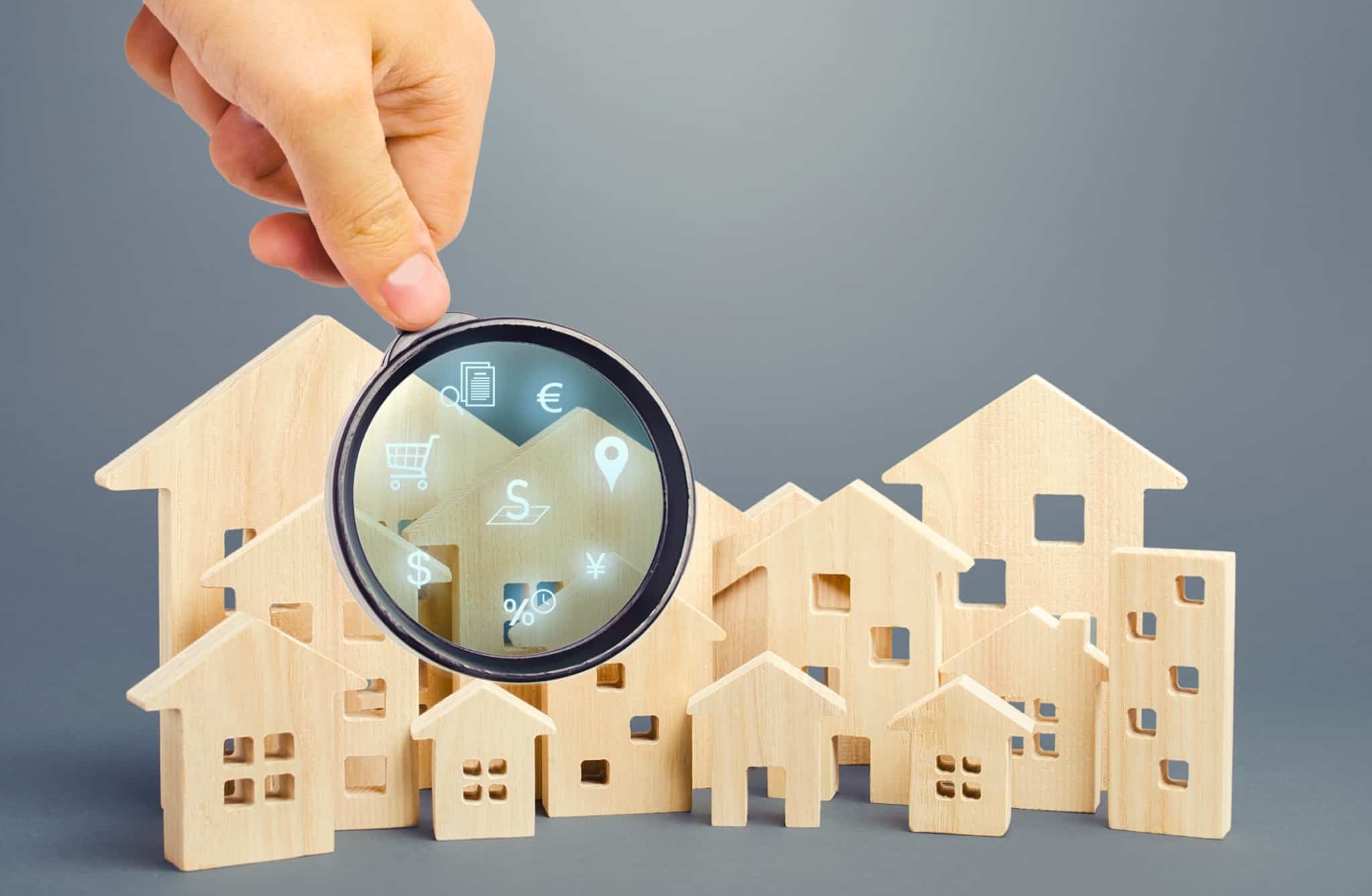 Figuras de casas e prédios em madeira e uma lupa à frente através da qual se veem simbolos de dinheiro, localização e compras