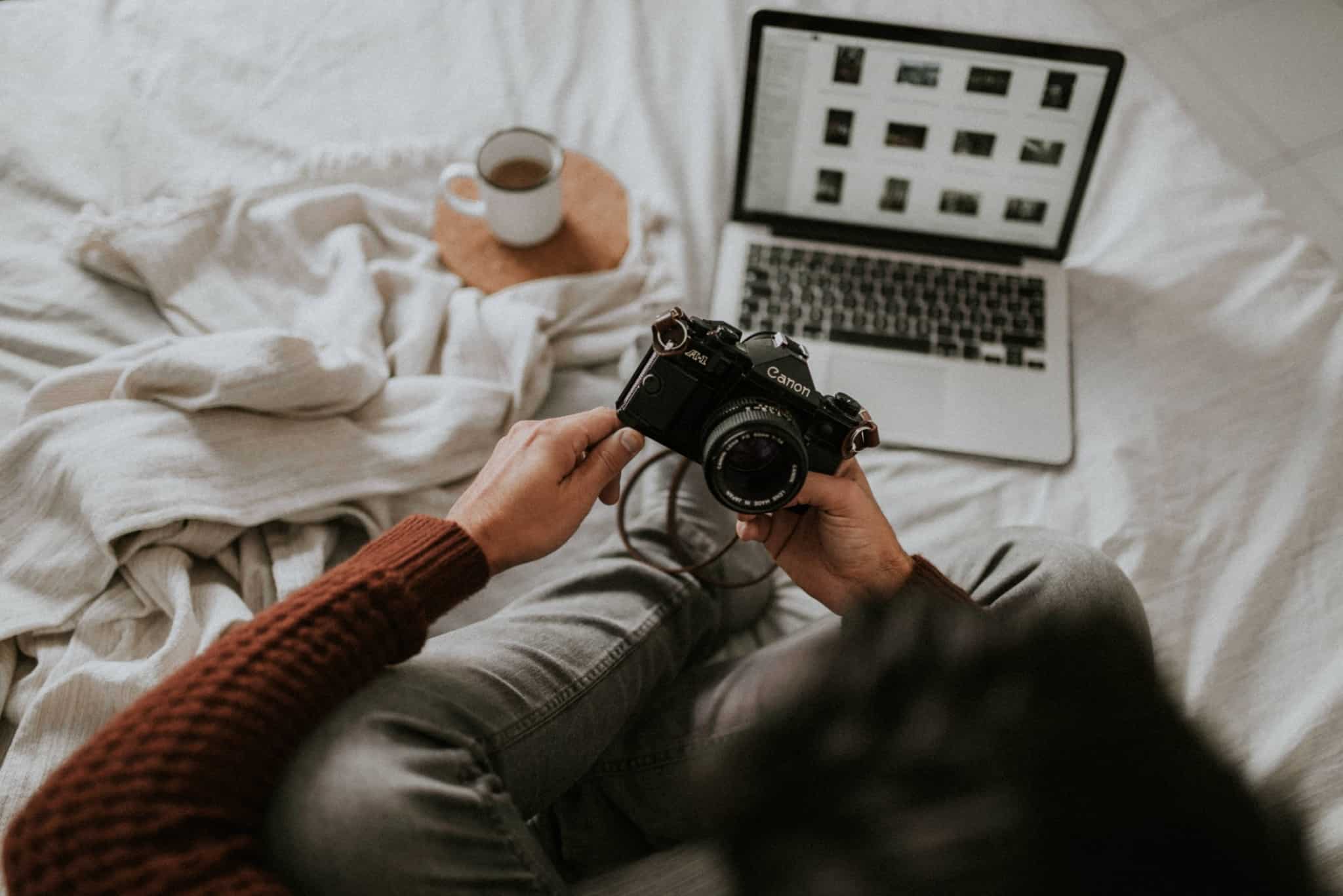 Pessoa com uma máquina fotográfica na mão. Á frente tem um computador, onde descarregou fotografias