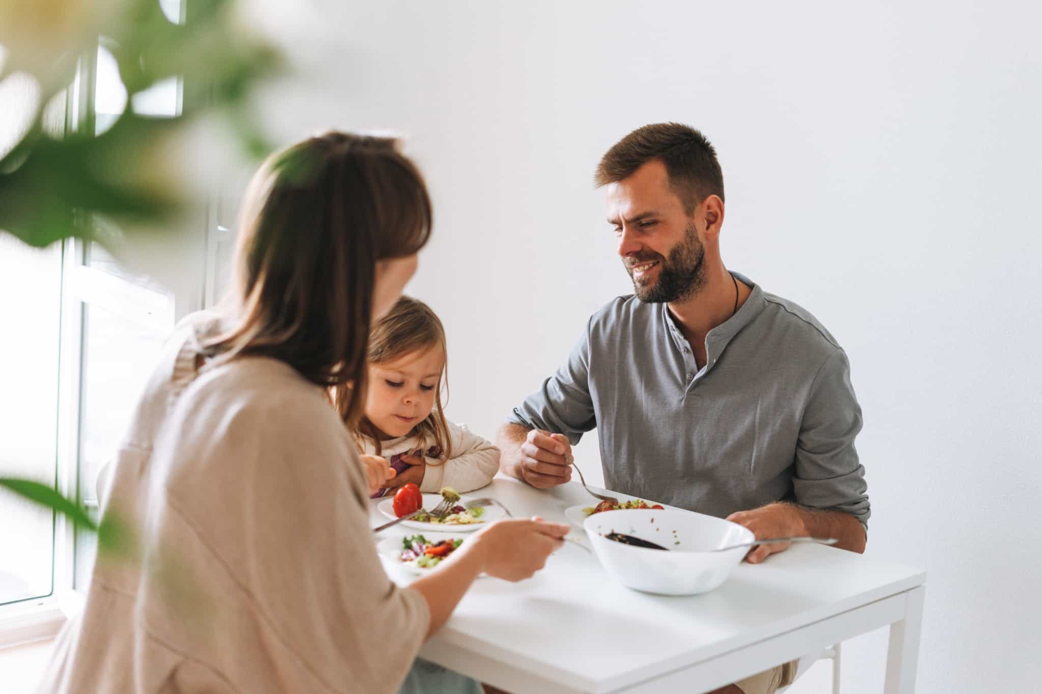 jovem casal sorridente, homem e mulher, sentados à mesa a fazer uma refeição com a sua filha pequena