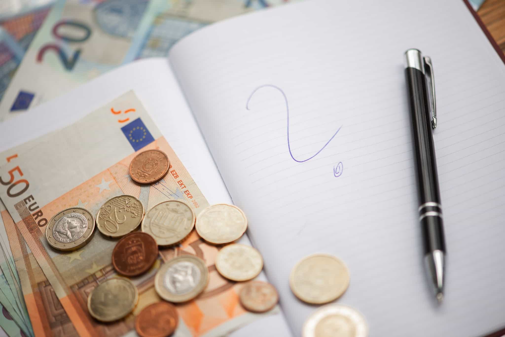 políticas monetárias: conceitos ilustrados por produção fotográfica com notas e moedas de euro, espalhados num caderno onde está um ponto de interrogação, escrito pela caneta que está pousada nesse caderno.