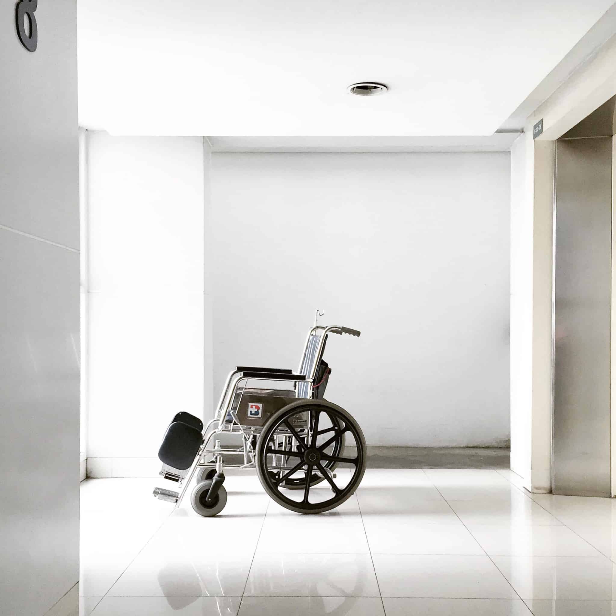 cadeira de rodas, vazia, parada no meio de um corredor também vazio, branco