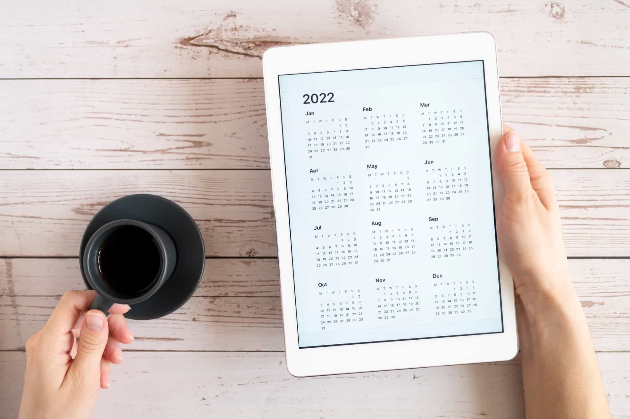Imagem de umas mãos a segurar num tablet com um calendário de 2022 e numa chávena de café - prazos máximos crédito habitação