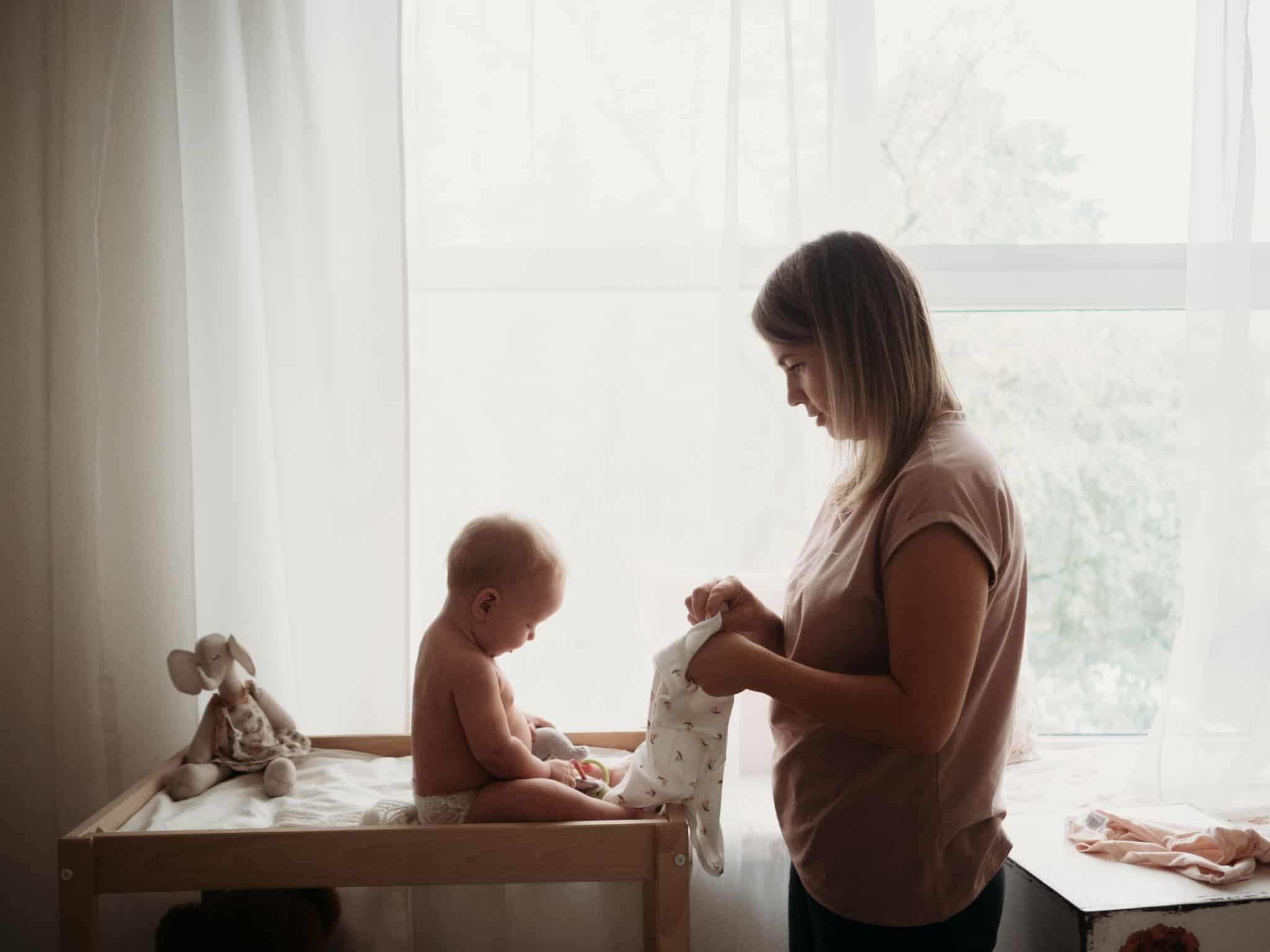 jovem mãe veste o seu bebé, no seu quarto, rodeado de todos os bens essenciais aos seus cuidados a ilustrar o direito ao subsídio parental em 2017