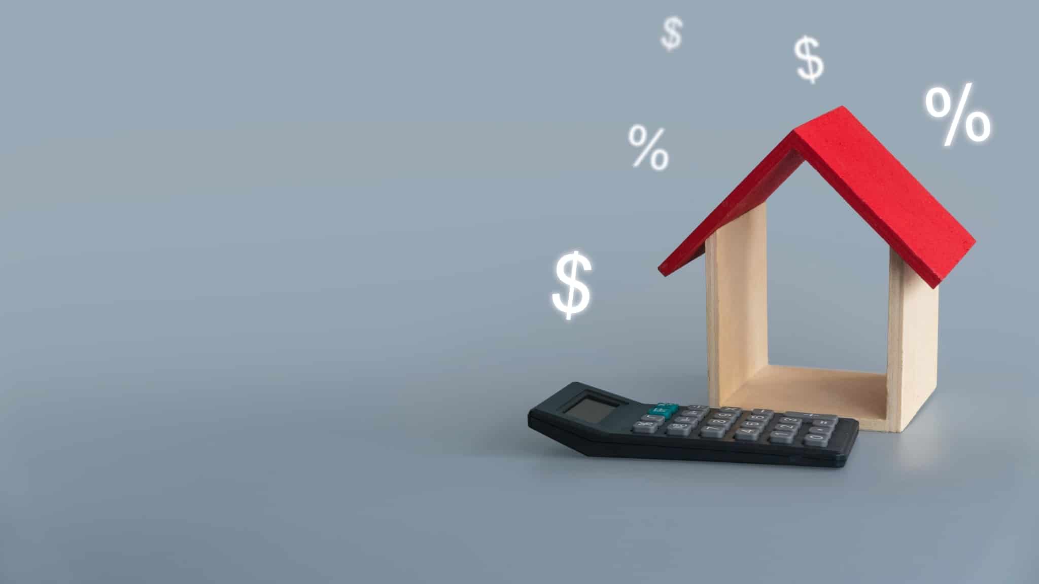 produção fotográfica mostra uma maqueta de uma casa, só com telhado vermelho e paredes laterais com calculadora ao pe e símbolos financeiros ao redor