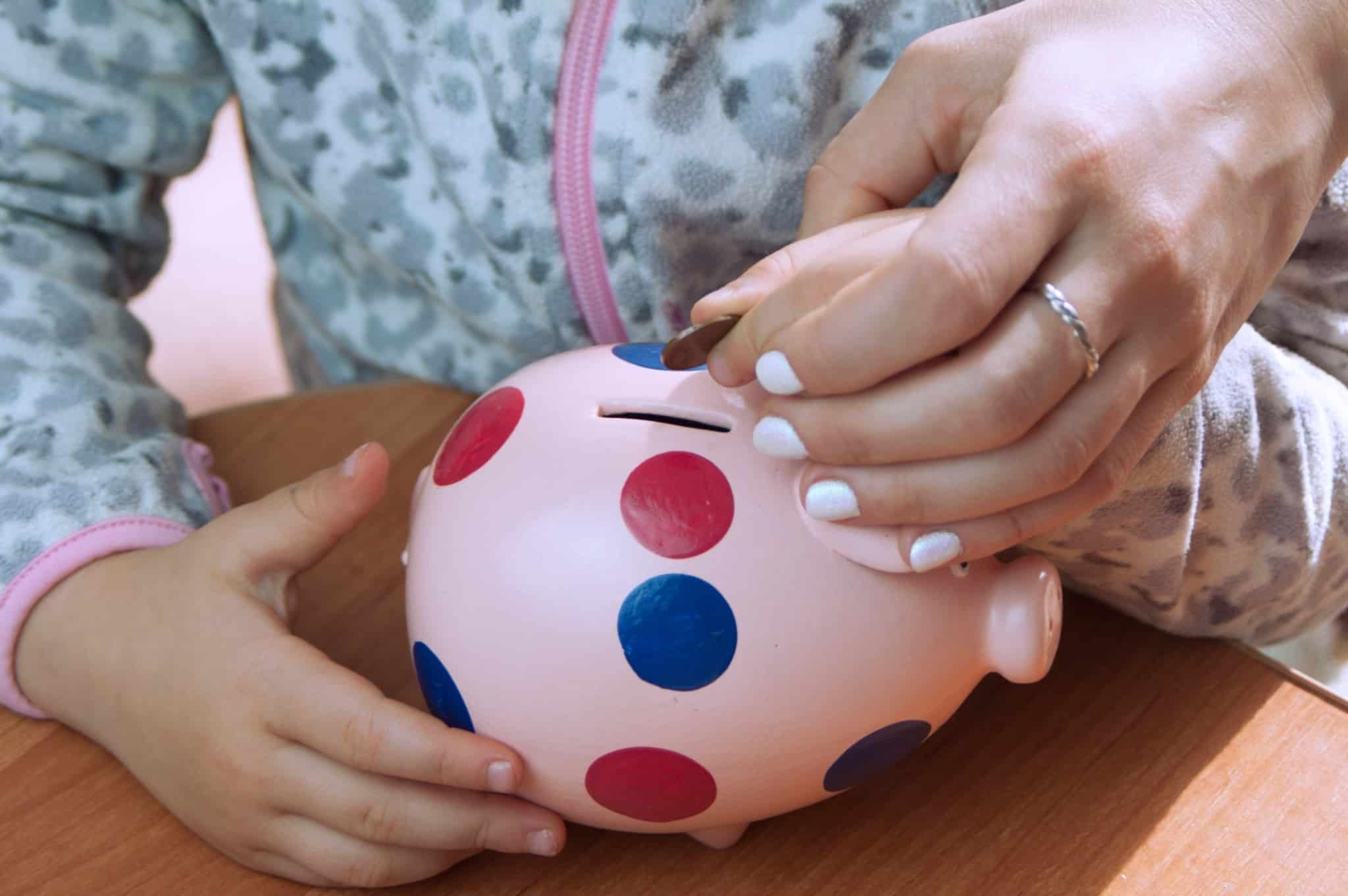 mãos de uma criança seguram um porquinho mealheiro, rosa e às bolinhas coloridas, apoiadas pela mão da mãe