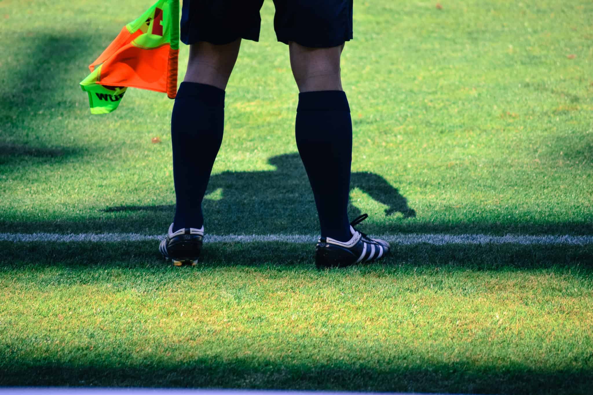 imagem de árbitro, apoiado por uma Bolsa de formação desportiva, junto á linha que delimita o campo de futebol, seguando uma bandeirola colorida