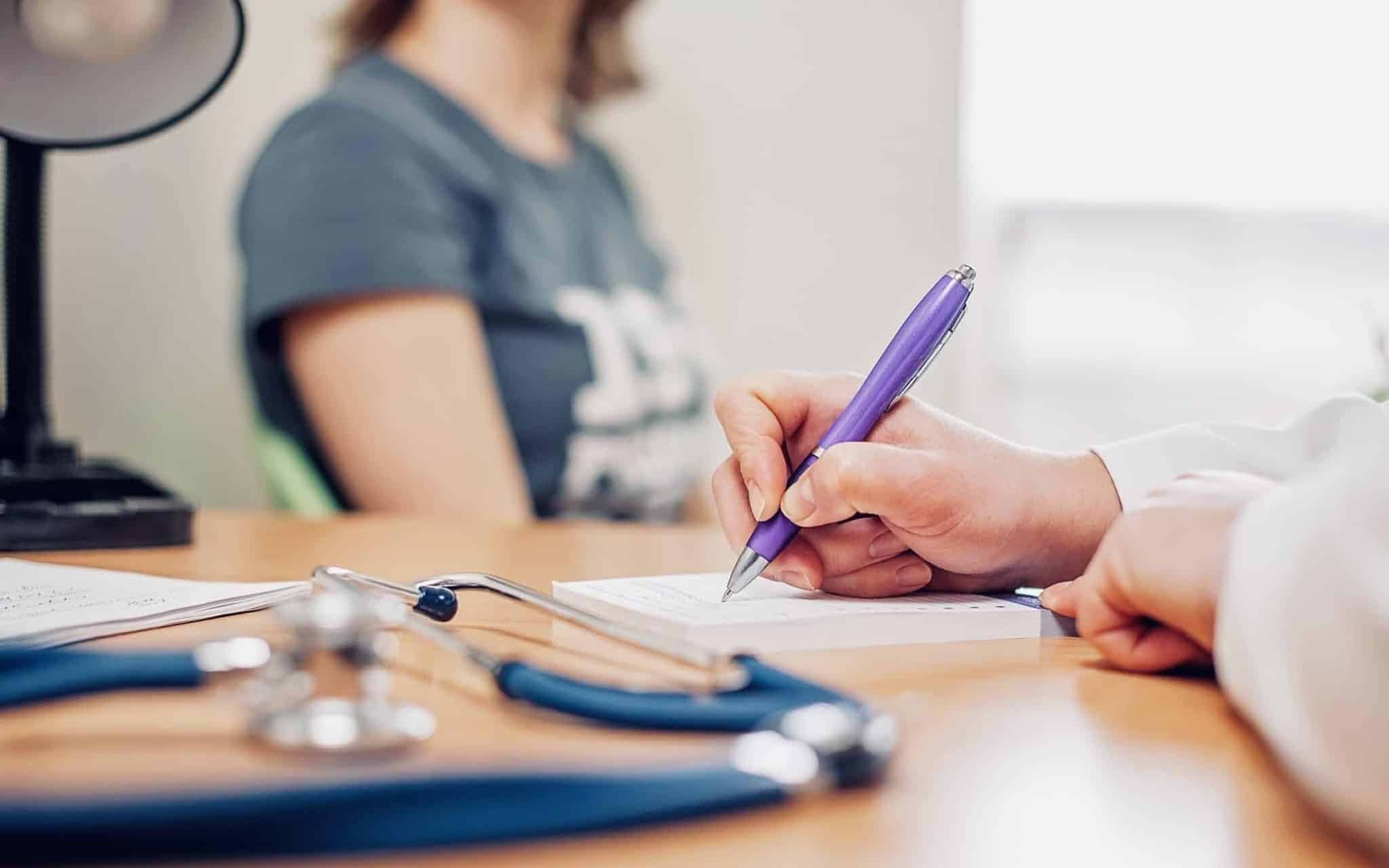 jovem mulher em consulta médica aguarda a prescrição que o médico está a redigir, a ilustrar a necessidade de recorrer a um simulador de seguro de saúde para reduzir os encargos com consultas e exames