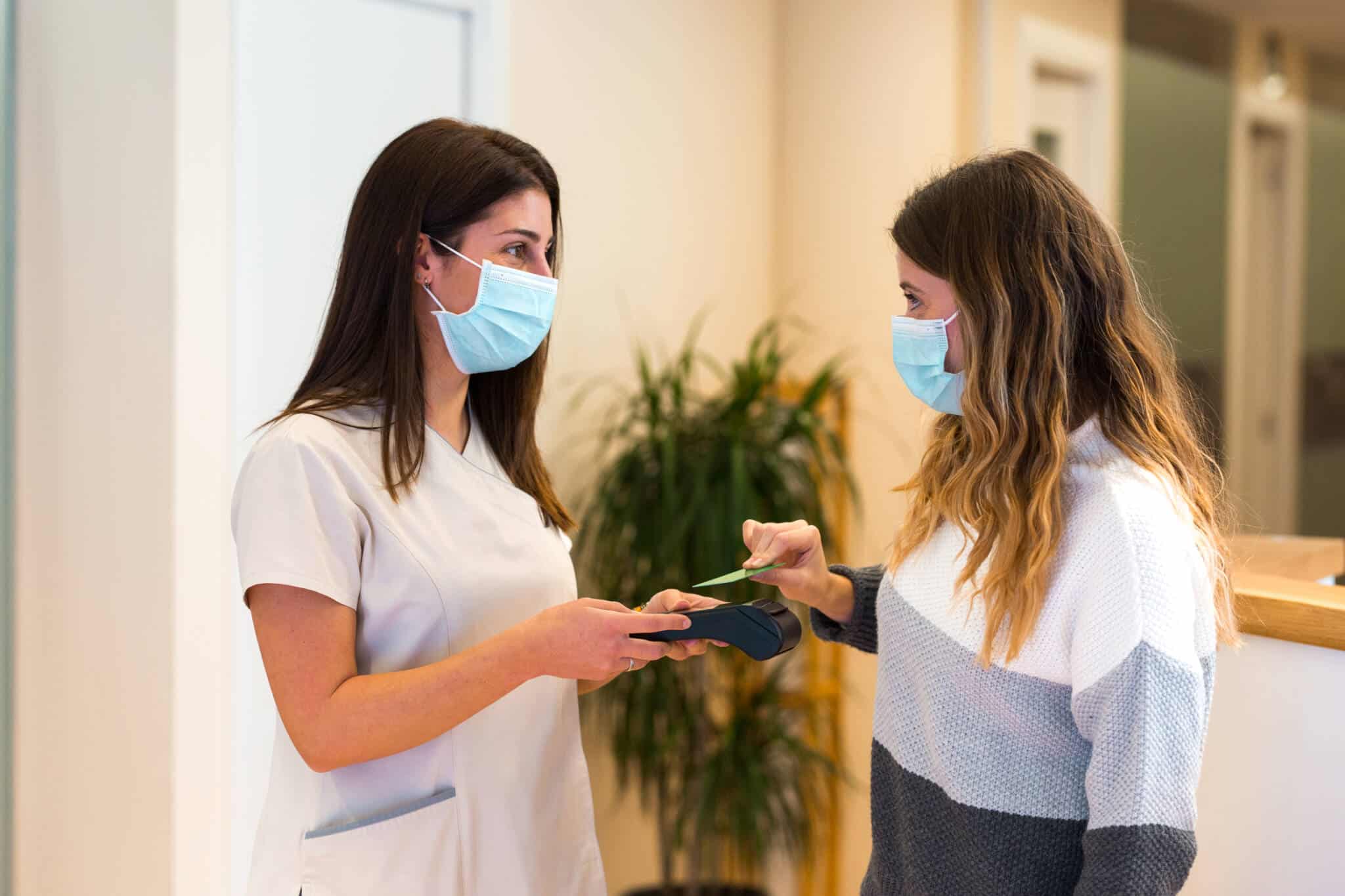 Enfermeira com máscara com multibanco na mão e paciente mulher de máscara a pagar com cartão bancário após intervenções cirúrgicas