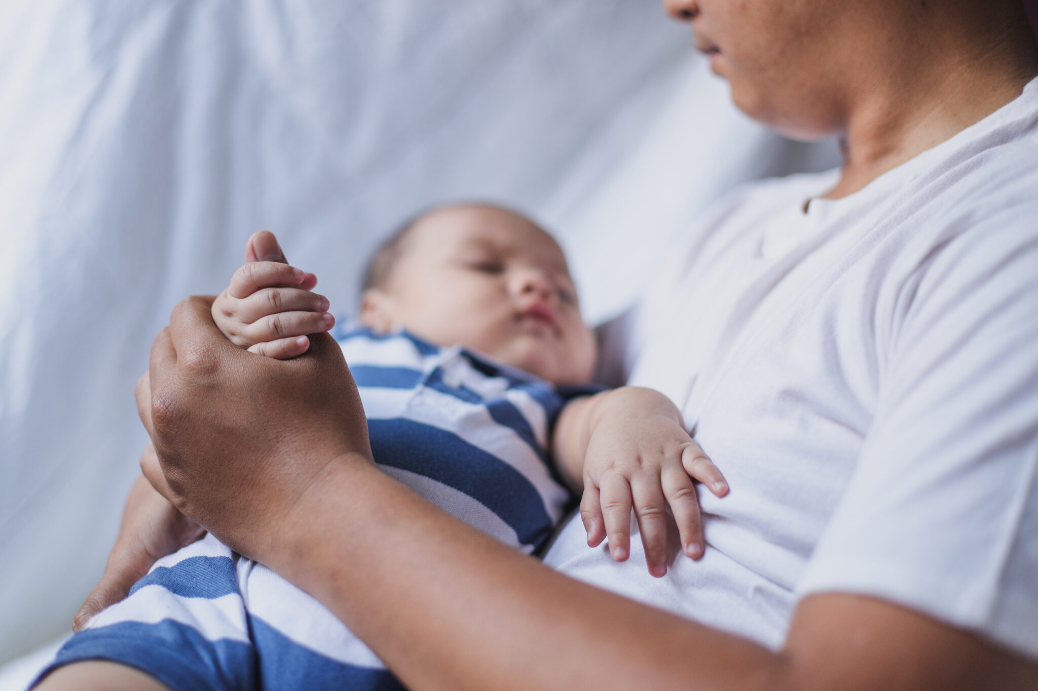 jovem pai segura o seu bebé nos braços, segurando a sua pequena mão, sentados no sofa branco, ilustrando as alterações na licença parental
