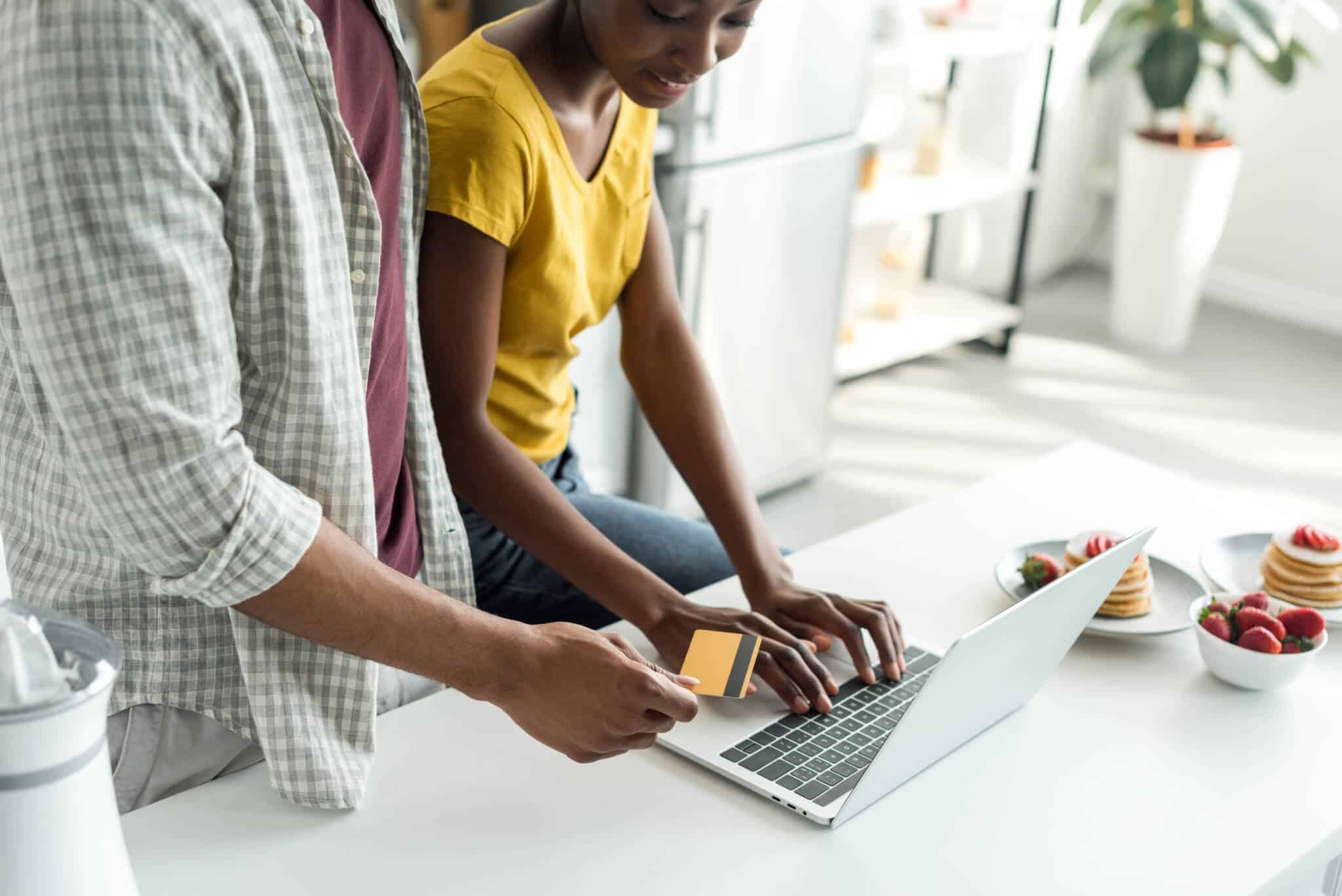 pai e filha adolescente na bancada da cozinha estão a consultar computador portátil para comparar preços online, e pagar com o cartão que estão a segurar