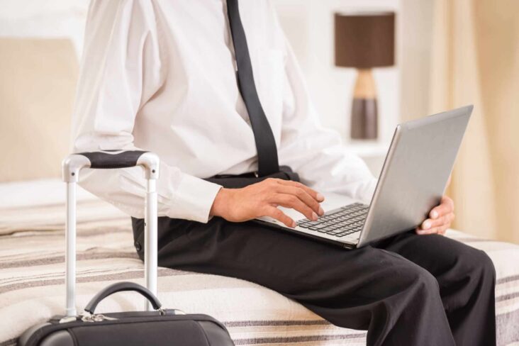 jovem homem sentado à beira da cama de hotel a consultar o computador portátil com a mala de viagem ao lado, a passar por fase laboral de mobilidade funcional