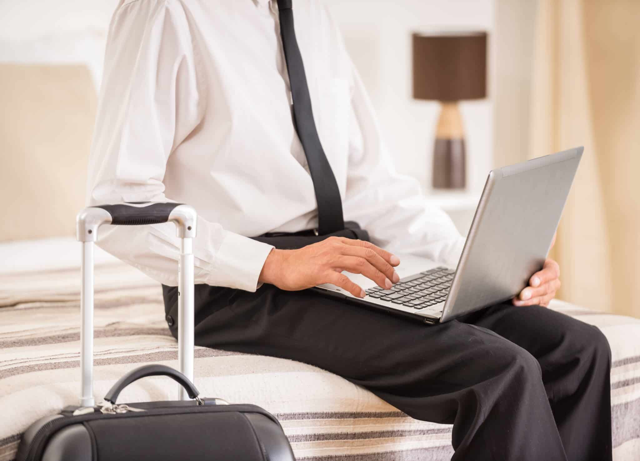 jovem homem sentado à beira da cama de hotel a consultar o computador portátil com a mala de viagem ao lado, a passar por fase laboral de mobilidade funcional