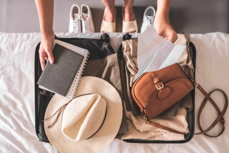 mala de viagem aberta, em cima da cama, enquanto jovem mulher prepara roupas e afins, contemplando seguro de viagem