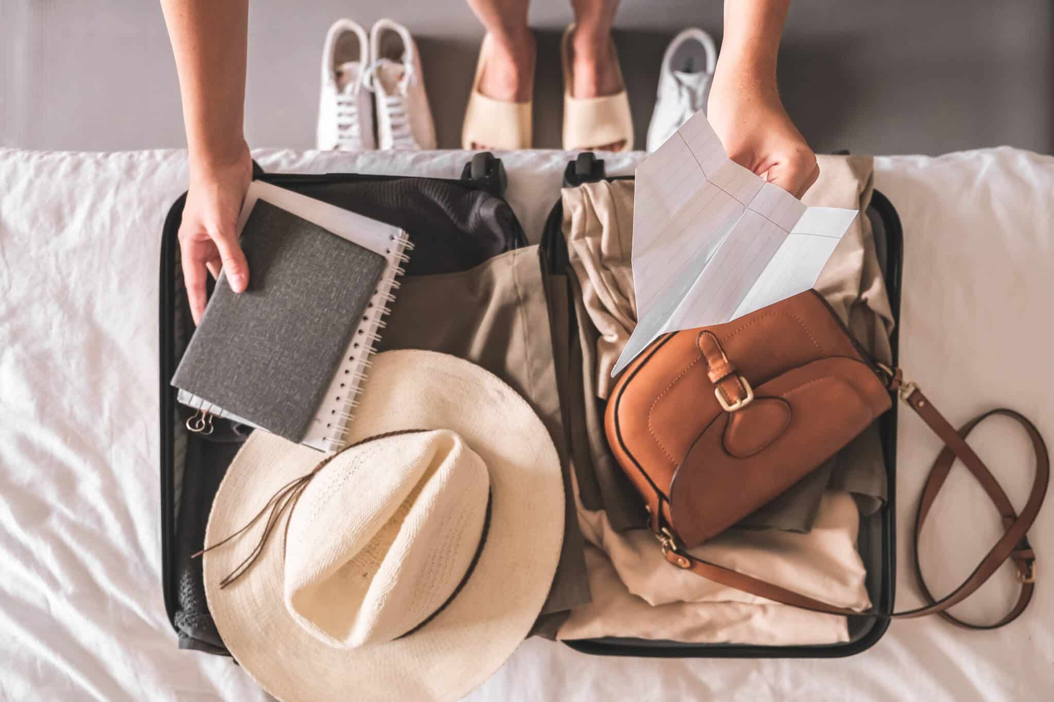 mala de viagem aberta, em cima da cama, enquanto jovem mulher prepara roupas e afins, contemplando seguro de viagem