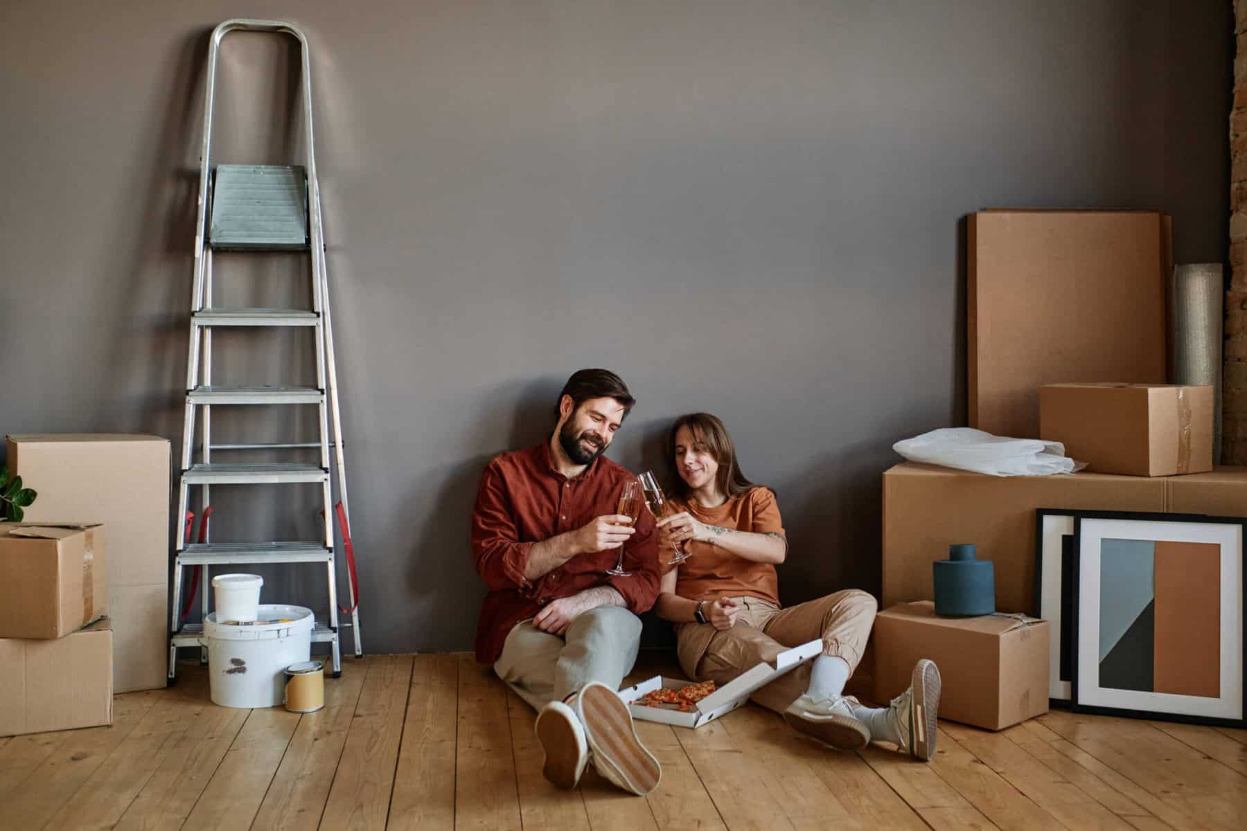 Casal sentado no chão, rodeado de caixas de cartão e material de construção, brindam com copos de champanhe a sua nova casa